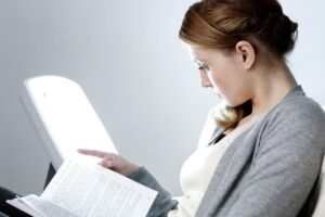 A young woman with seasonal affective disorder (SAD) sits in front of a bright SAD light therapy lamp while she reads a book