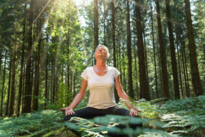 A woman sits cross-legged relaxed in the forest practicing deep breathing or meditation - Forest Bathing - A Natural Remedy for Better Sleep - by Simply Good Sleep